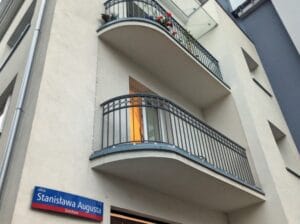 Beżowa, drobna siatka zamontowana na balkonie na pierwszym piętrze na warszawskim Grochowie. Siatka jest mało widoczna na tle białego bloku.