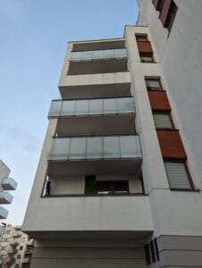 Widok z chodnika na pion balkonów w budynku wielorodzinnym. Balkon na drugim piętrze z zamontowaną siatką beżową. Siatka na drugim piętrze na warszawskim osiedlu zupełnie niewidoczna z poziomu chodnika.