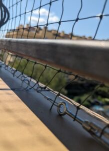 Metoda instalacji siatki na balkon. Na zdjęciu widać wywiercony otwór z zamontowanym kołkiem i hakiem do którego zamontowana jest linka stalowa, do której doszyta jest siatka.