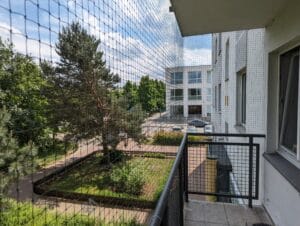 Siatka na balkon zamontowana metodą inwazyjną. Siatka wzmocniona drucikiem w kolorze oliwkowym. W tle za siatką słoneczny dzień i zielone drzewa.