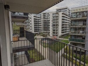 Wytrzymała, gruba siatka na balkon w kolorze beżowym z oczkiem 40x40 mm, zamontowana bezinwazyjnie po wewnętrznej stronie barierki. Instalacja wykonana na czwartym piętrze na warszawskim Żoliborzu.